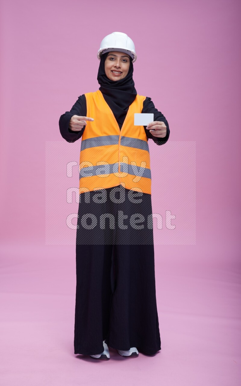 Saudi woman wearing Abaya with engineer vest and helmet standing holding ATM card on pink background