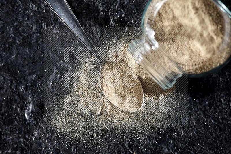 A flipped herbal glass jar and a metal spoon full of white pepper powder with spilled powder on textured black flooring