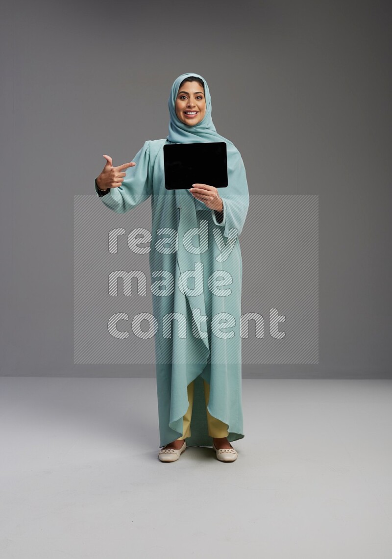 Saudi Woman wearing Abaya standing showing tablet to camera on Gray background