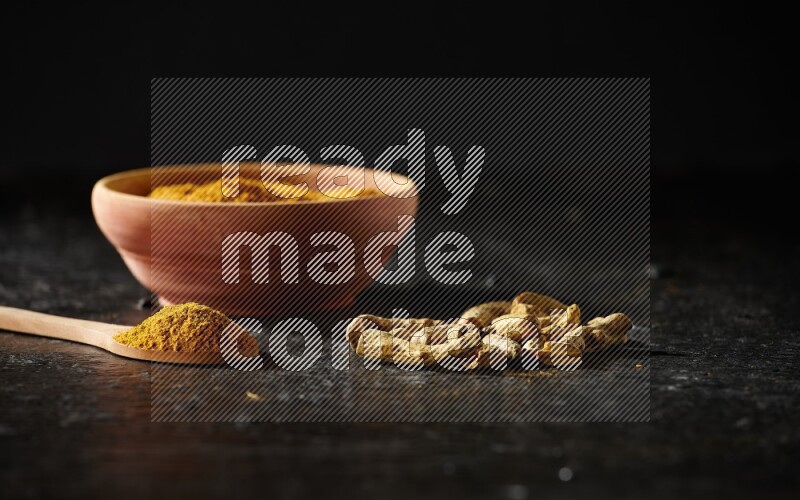 A wooden bowl and a wooden spoon full of turmeric powder with dried turmeric fingers on textured black flooring