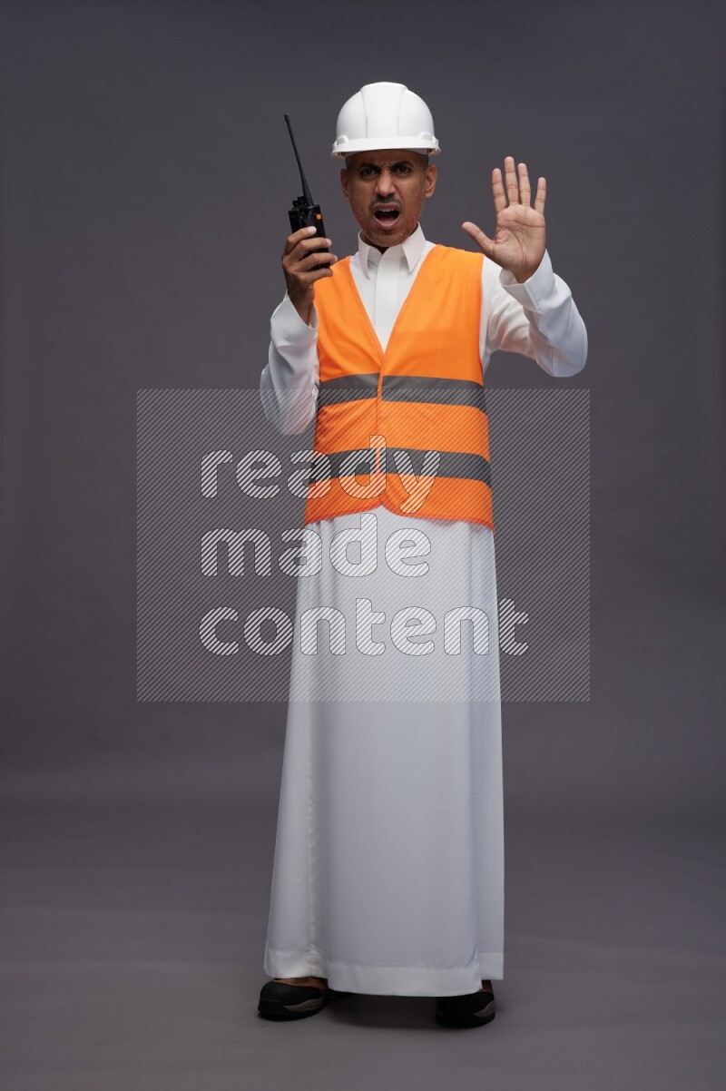 Saudi man wearing thob with engineer vest standing holding walkie-talkie on gray background