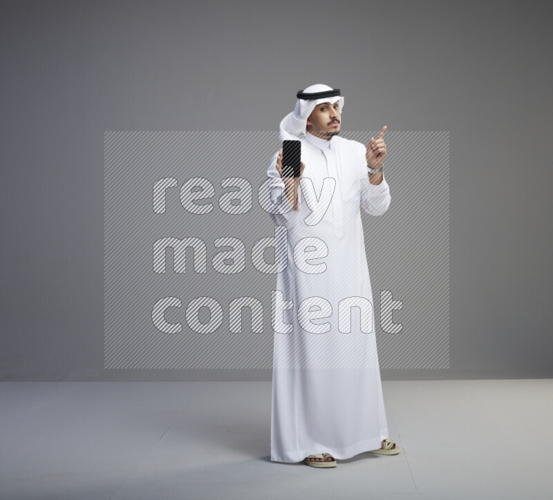 A Saudi man standing wearing thob and white shomag showing phone to camera on gray background