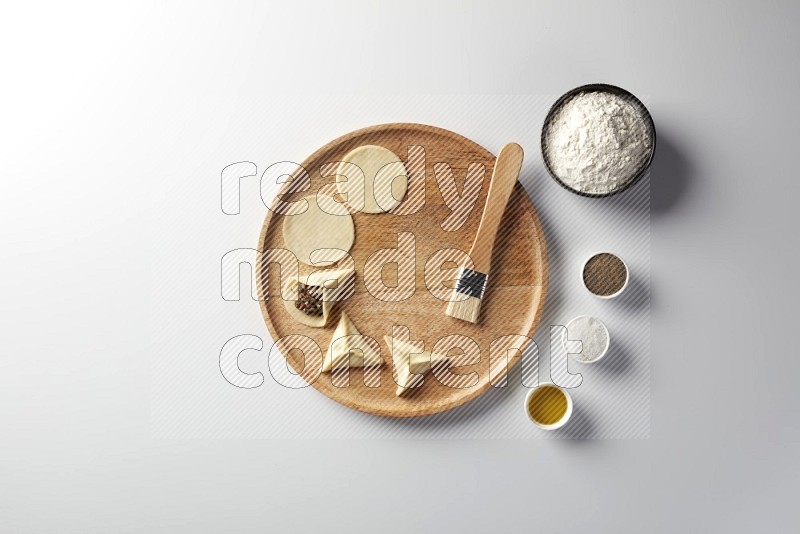 two closed sambosas and one open sambosa filled with meat while flour, salt, black pepper and oil with oil brush aside in a wooden dish on a white background