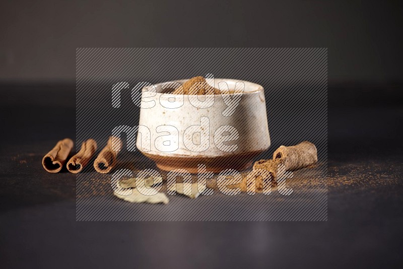 بودرة القرفة في وعاء فخاري أبيض وأعواد القرفة وأوراق الغار على خلفية سوداء