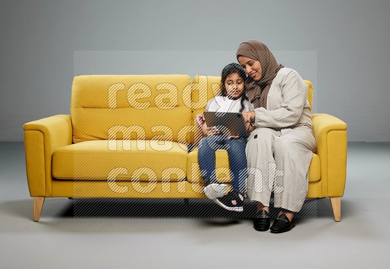 A girl with her mother on yellow sofa and watching on iPad on gray background
