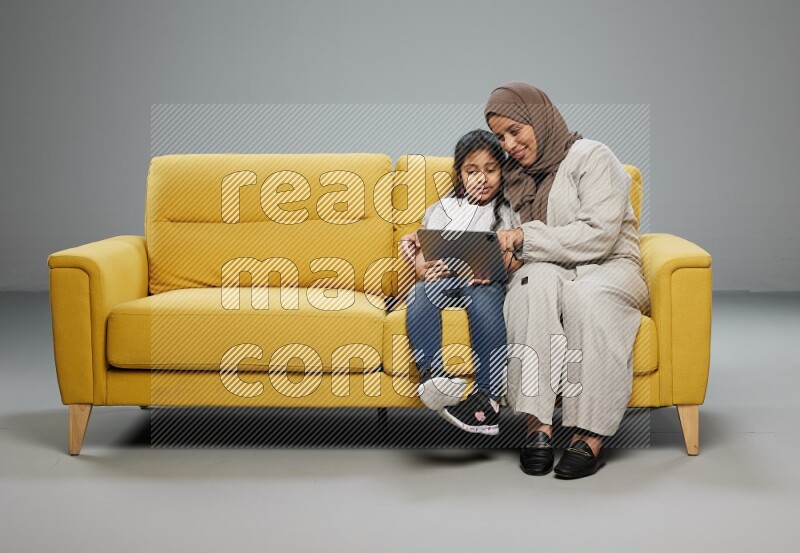 A girl with her mother on yellow sofa and watching on iPad on gray background