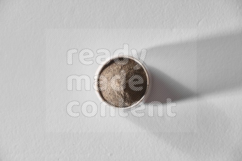 A white ceramic bowl full of black pepper powder and black pepper beads spread on white flooring