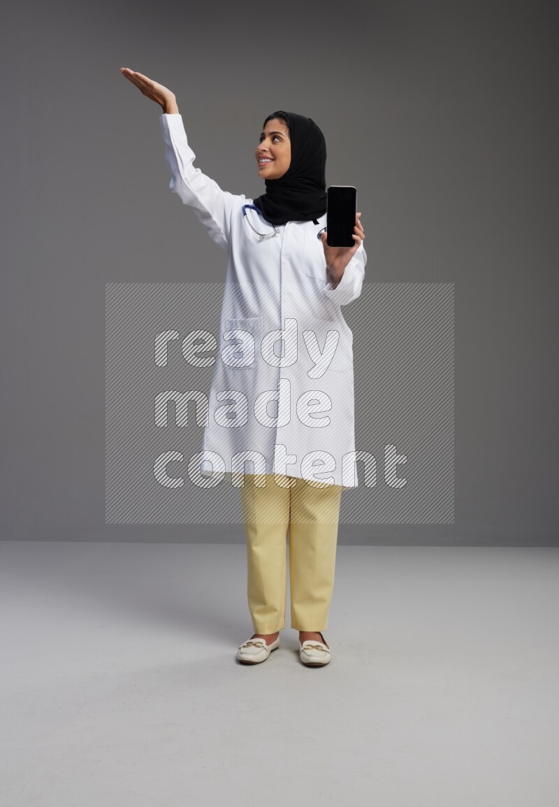 Saudi woman wearing lab coat with stethoscope standing showing phone to camera with sign in the back on Gray background