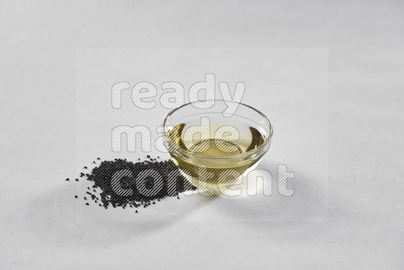 وعاء زجاجي ممتلئ بزيت حبة البركة مع بذور حبة البركة بجانبه على أرضية بيضاء