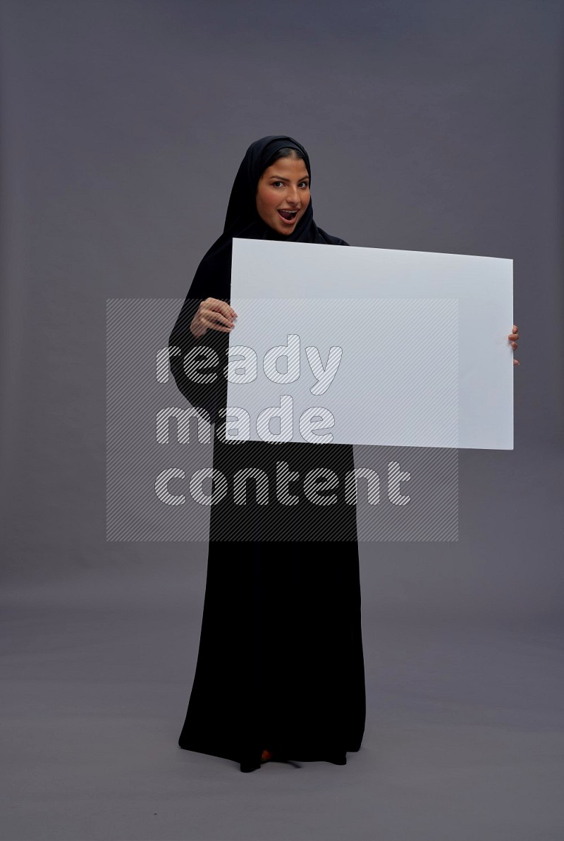 Saudi woman wearing Abaya standing holding white board on gray background