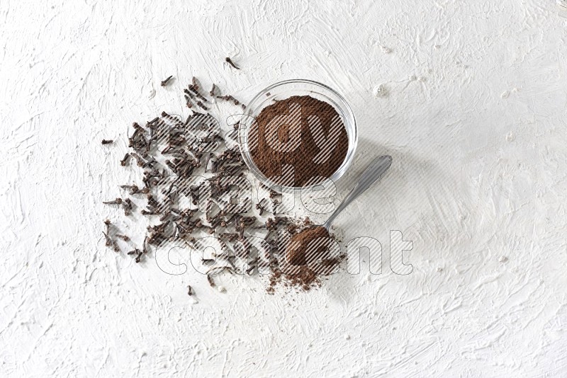 A glass bowl and a metal spoon full of cloves powder with cloves grains spread on a textured white flooring