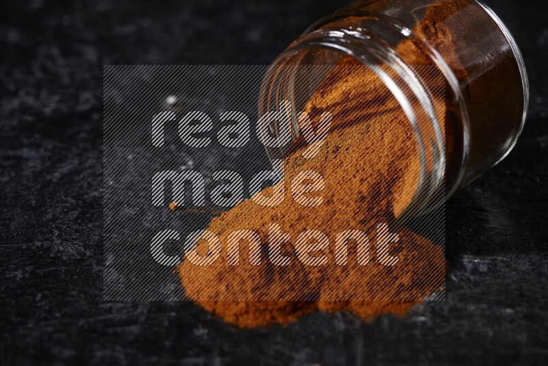 A glass jar full of ground paprika powder flipped with some spilling powder on black background