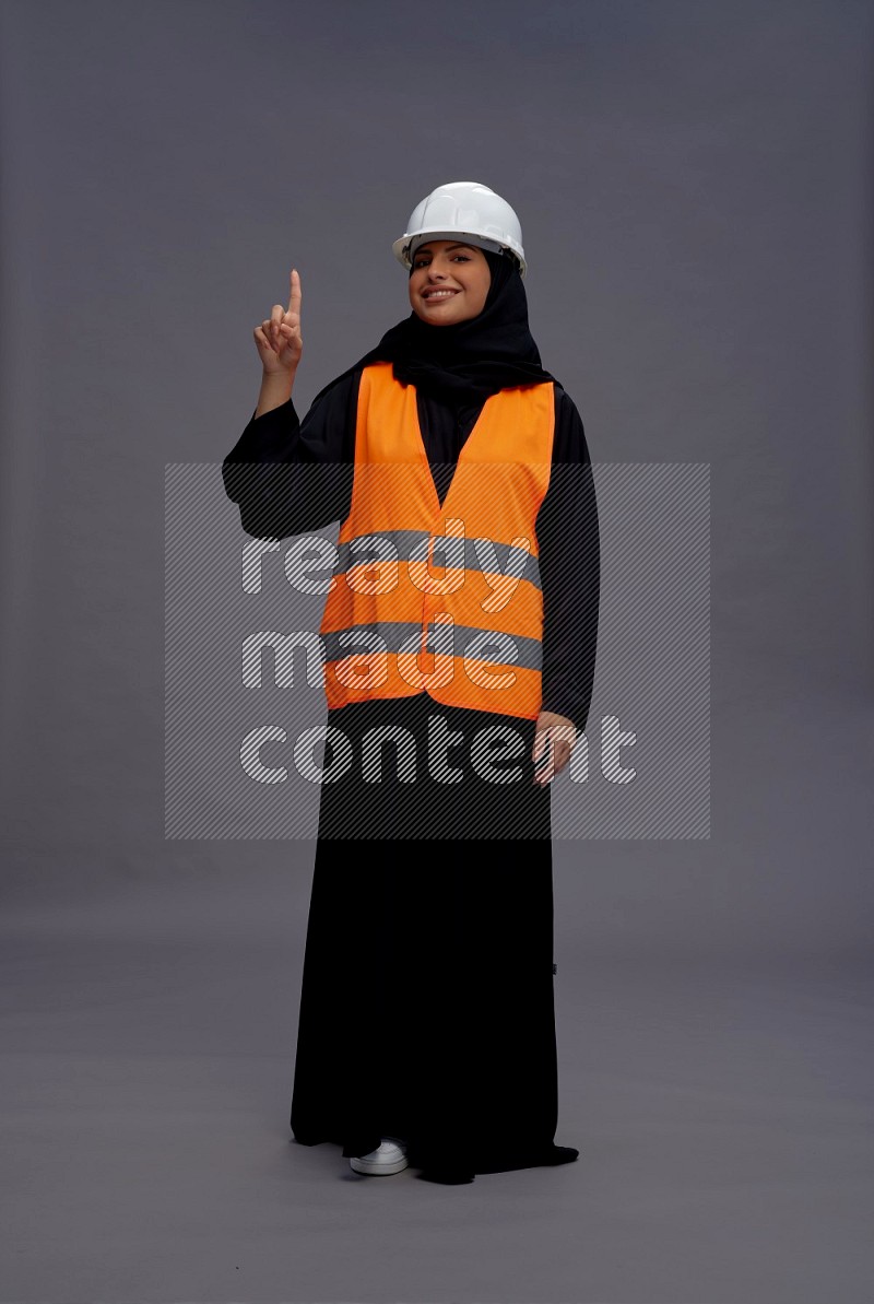 Saudi woman wearing Abaya with engineer vest standing interacting with the camera on gray background