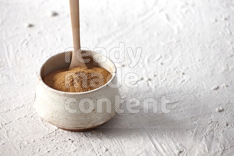 Ceramic beige bowl full of cinnamon powder with a wooden spoon on a textured white background