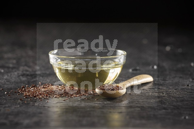 A glass bowl full of flaxseeds oil and wooden spoon full of flaxseeds with seeds spread on a textured black flooring