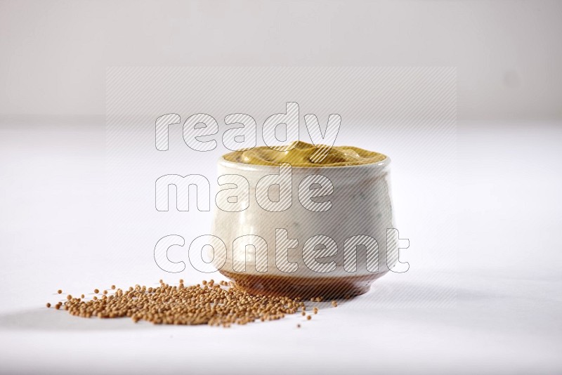 A beige pottery bowl full of mustard paste with mustard seeds underneath on white flooring