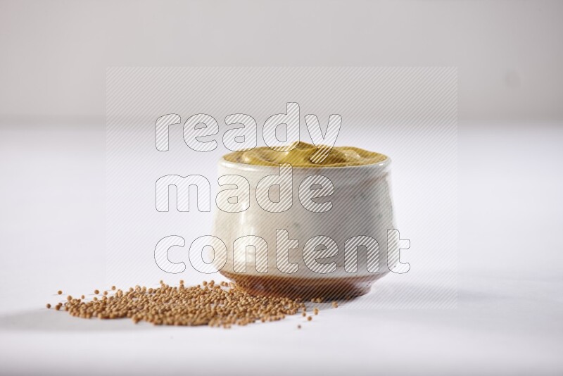 A beige pottery bowl full of mustard paste with mustard seeds underneath on white flooring