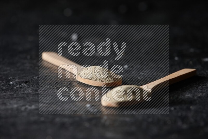 2 wooden spoons full of white pepper powder on textured black flooring