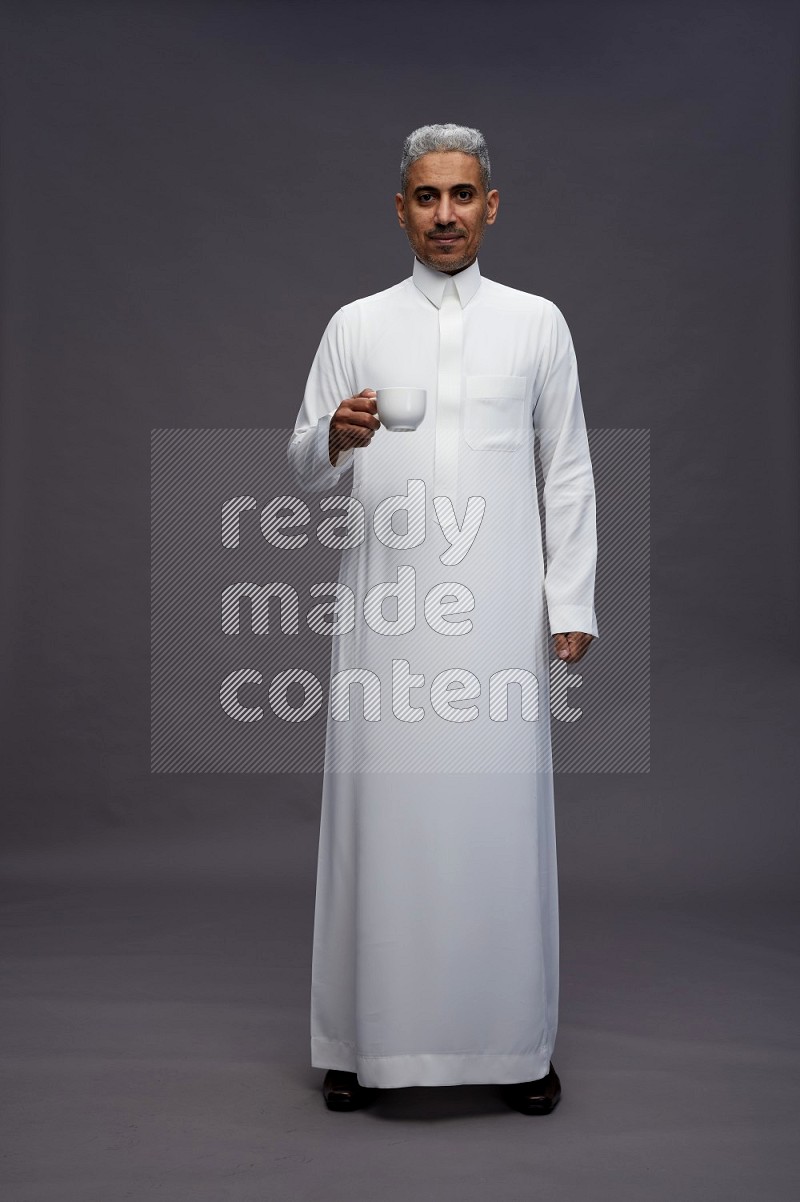 Saudi man wearing thob standing holding mug on gray background