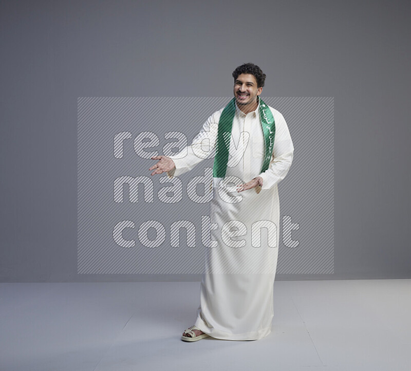 A Saudi man standing wearing thob and Saudi flag scarf on gray background