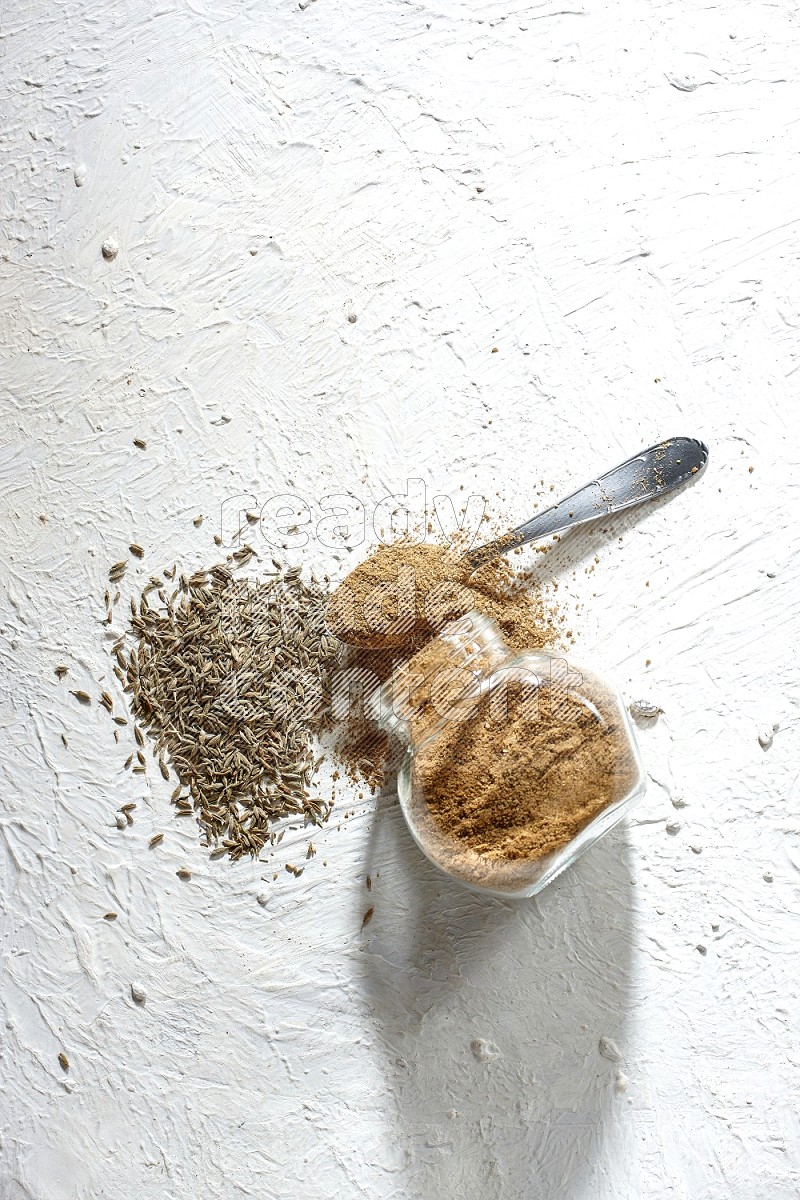 A glass spice jar and metal spoon full of cumin powder and the jar flipped and powder spilled out with cumin seeds on textured white flooring