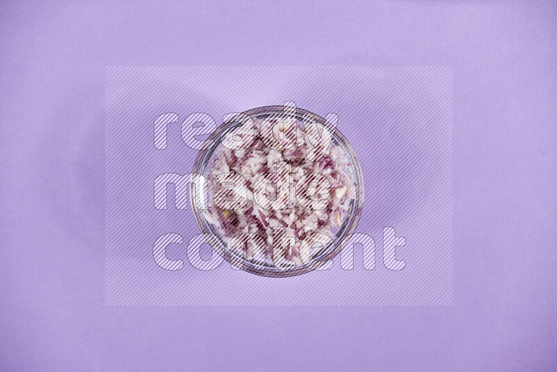 A glass bowl full of chopped red onions with halved onions beside it on purple background