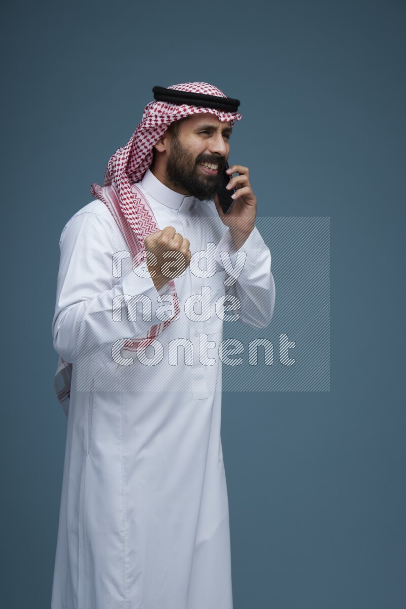 A man having a Call in a blue background wearing Saudi Thob with Shomag