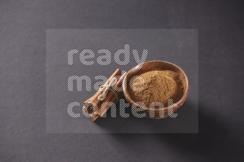 Cinnamon sticks stacked and bounded beside a wooden bowl full of cinnamon powder and a wooden spoon full of powder on black background