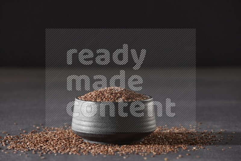 وعاء فخاري أسود ممتلئ بحبوب بذر الكتان محاط بالبذور على أرضية سوداء