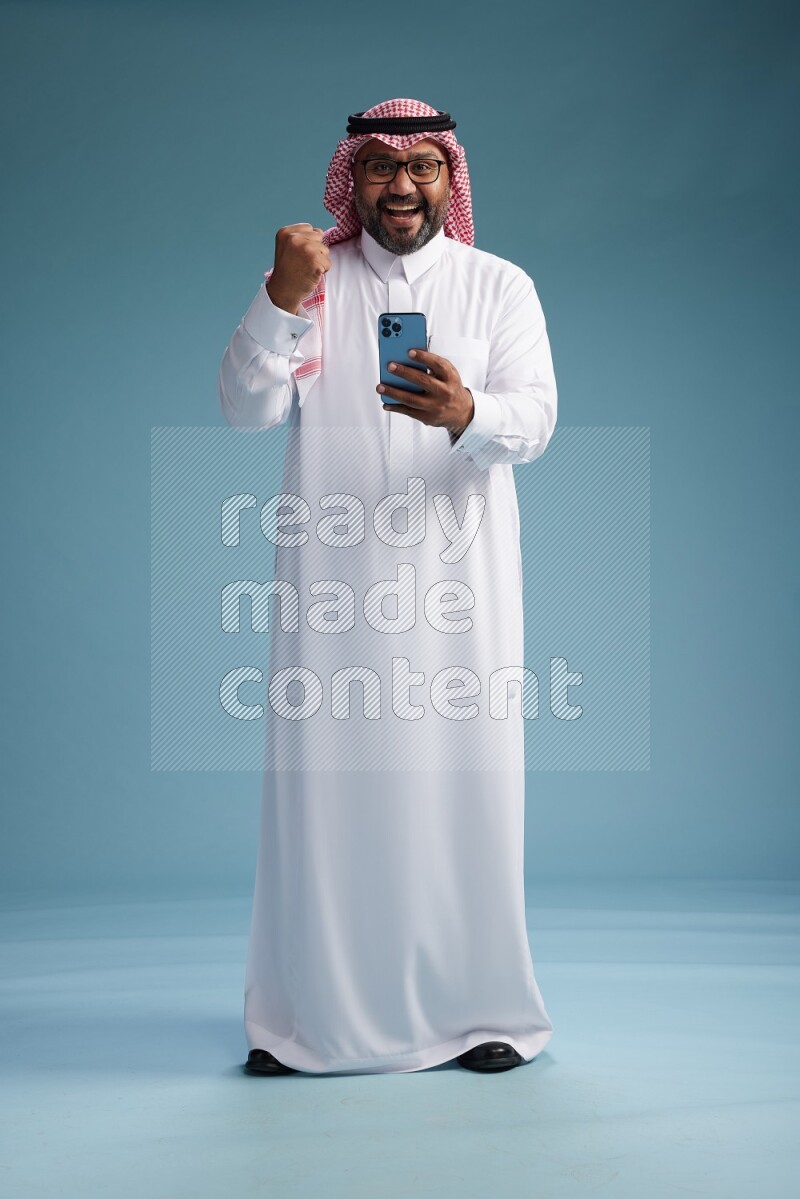 Saudi Man with shimag Standing texting on phone on blue background