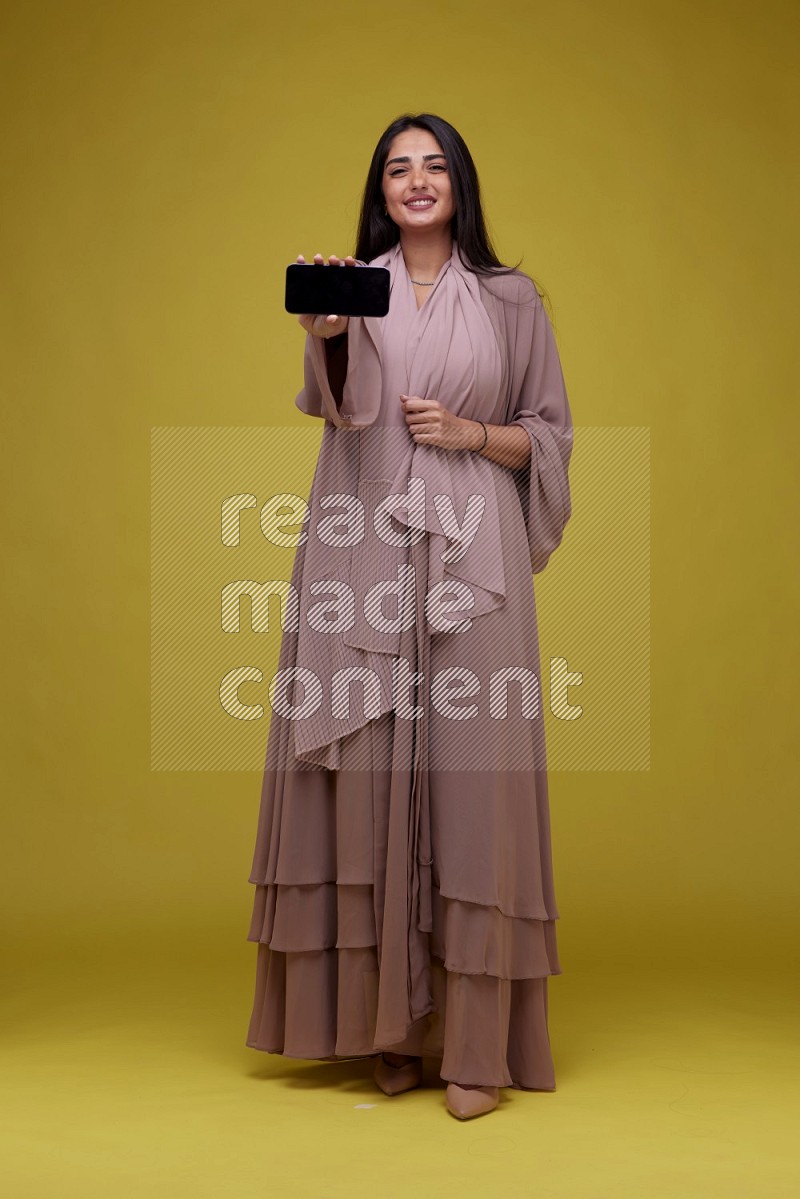 A woman Showing SmartPhone Screen on a Yellow Background wearing Brown Abaya