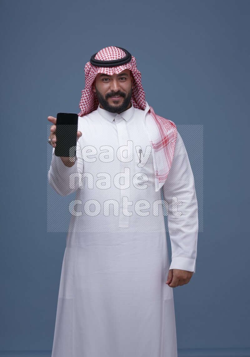 A man Showing his smartphone Screen on Blue Background wearing Saudi Thob