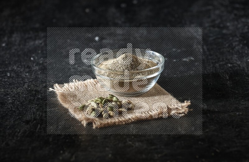 A glass bowl full of cardamom powder with cardamom seeds on a burlap piece on textured black flooring