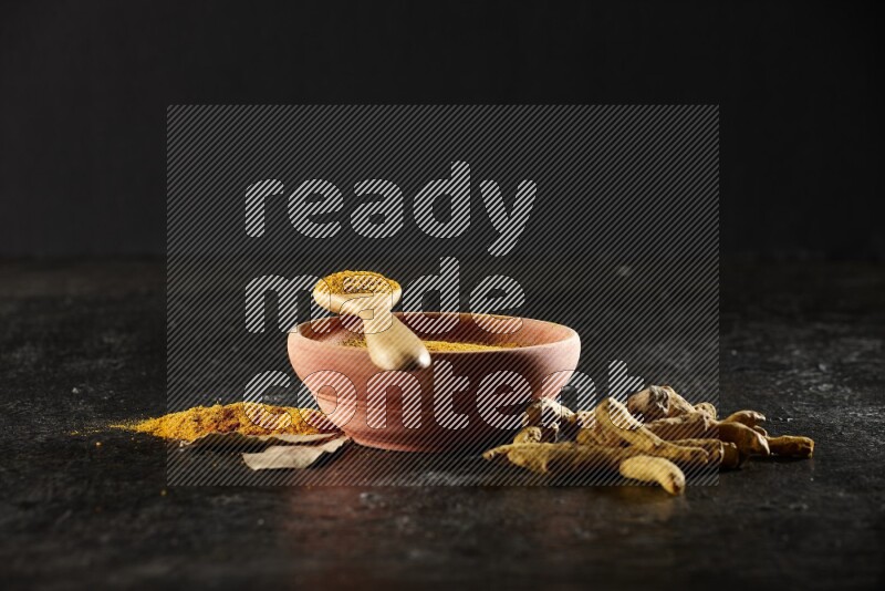 A wooden bowl and a wooden spoon full of turmeric powder with dried turmeric fingers on textured black flooring