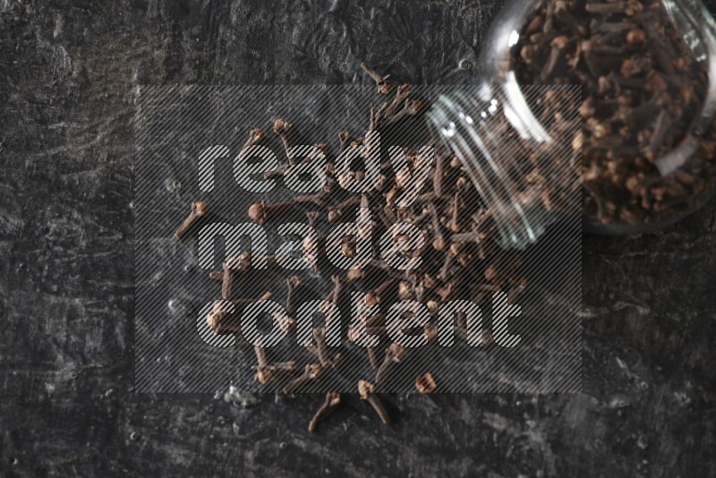 A flipped glass spice jar full of cloves and cloves came out of it on textured black flooring