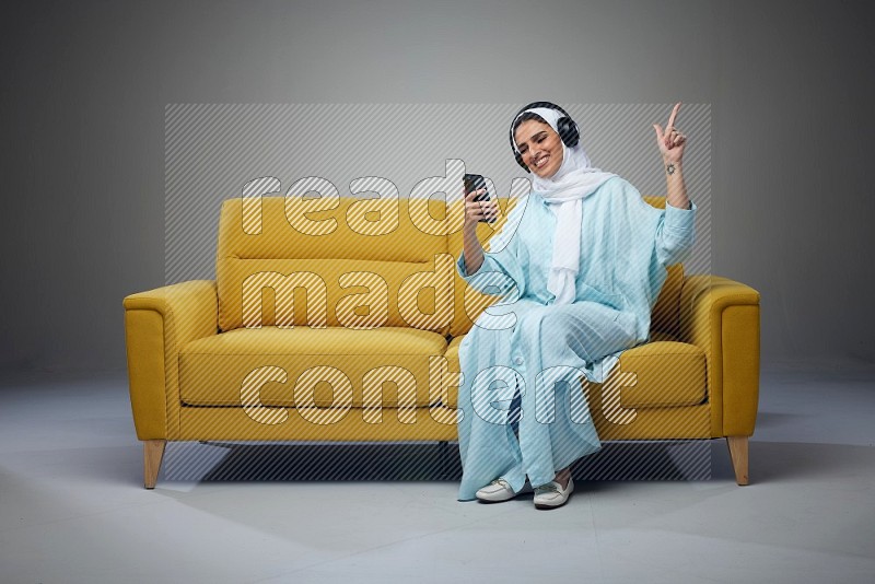 A Saudi woman wearing a light blue Abaya and a white head scarf sitting on a yellow sofa and holding an iPad while wearing headphone eye level on a grey background