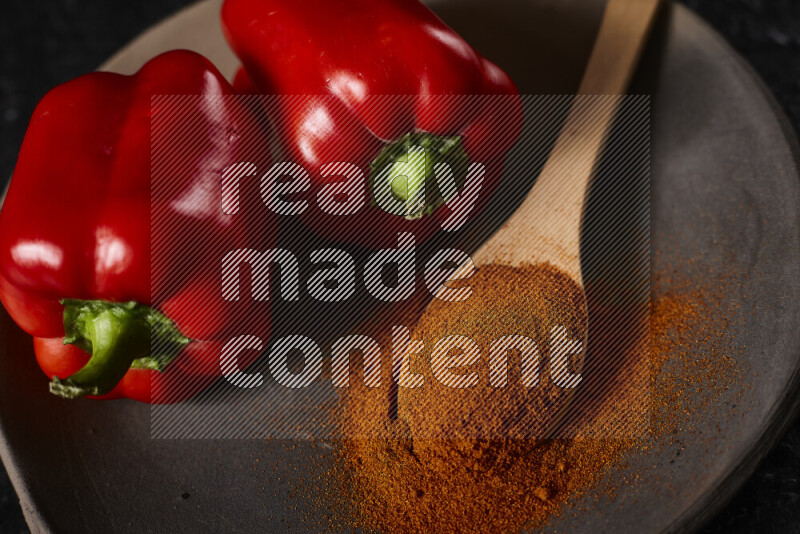 A wooden spoon full of ground paprika powder with red bell peppers, all on a pottery plate on black background