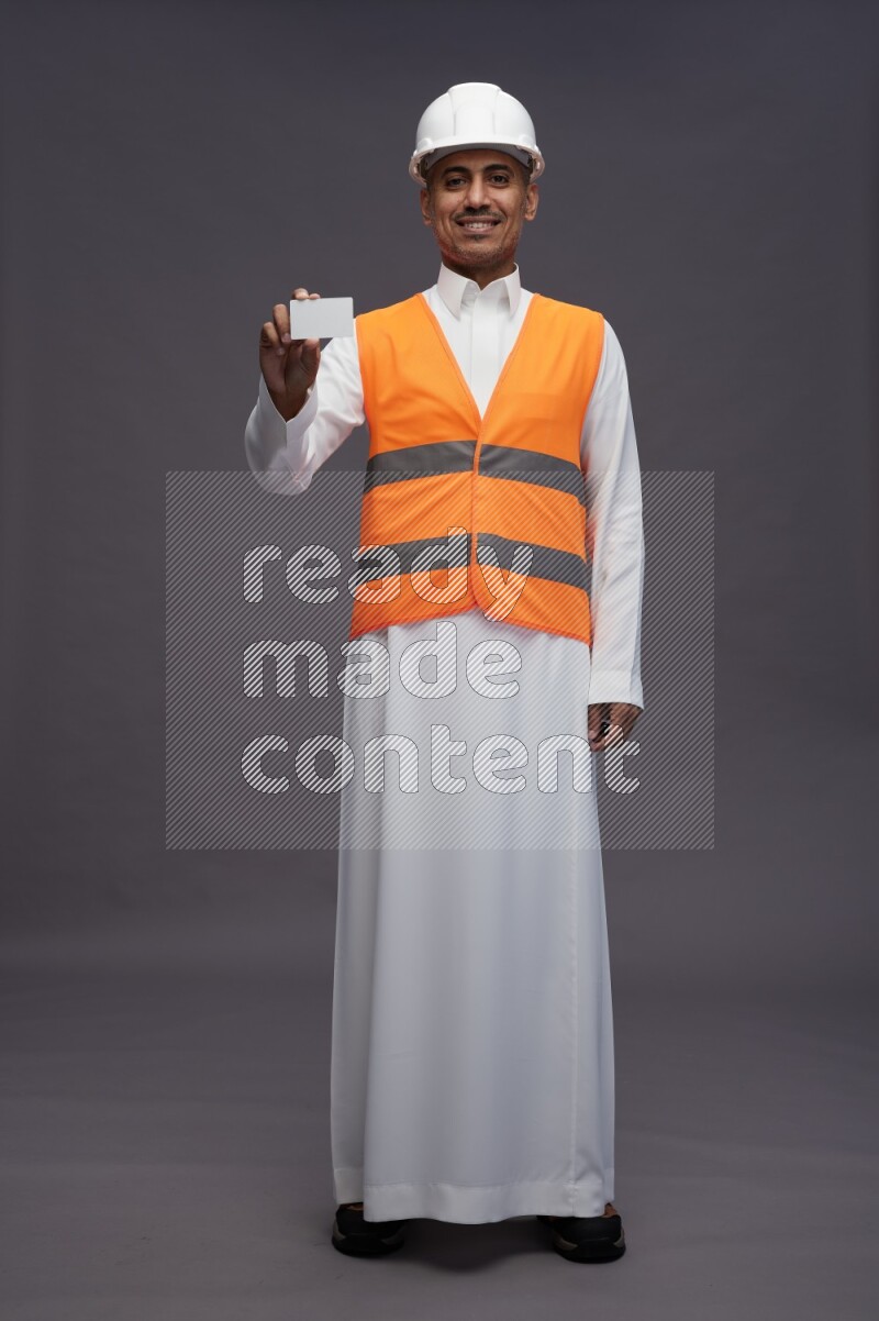 Saudi man wearing thob with engineer vest standing holding ATM card on gray background