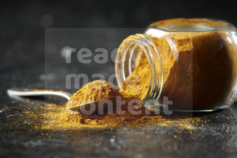 A flipped glass spice jar and a metal spoon full of turmeric powder and powder spilled out of it on textured black flooring