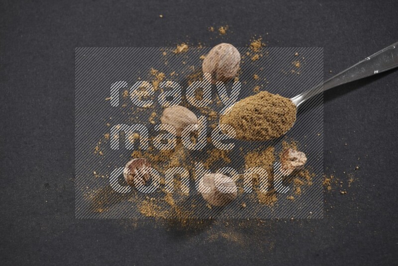 A metal spoon full of nutmeg powder with nutmeg powder and whole seeds beside it on a black flooring