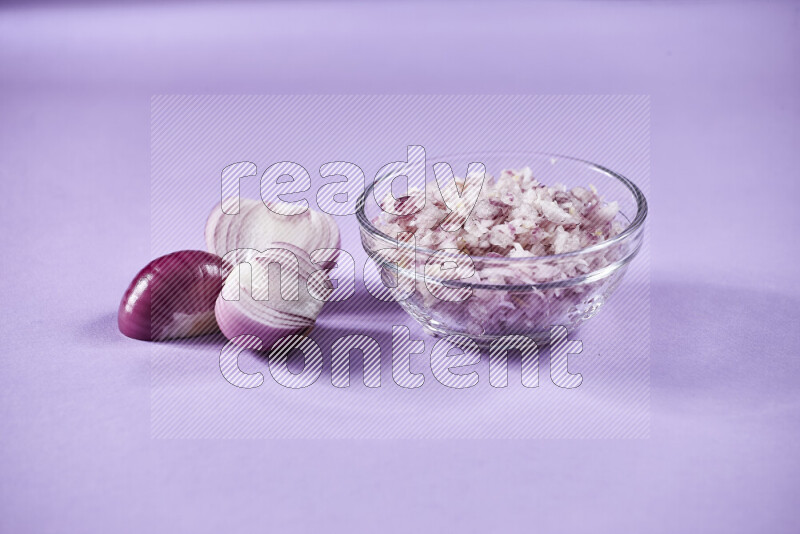 A glass bowl full of chopped red onions with halved onions beside it on purple background