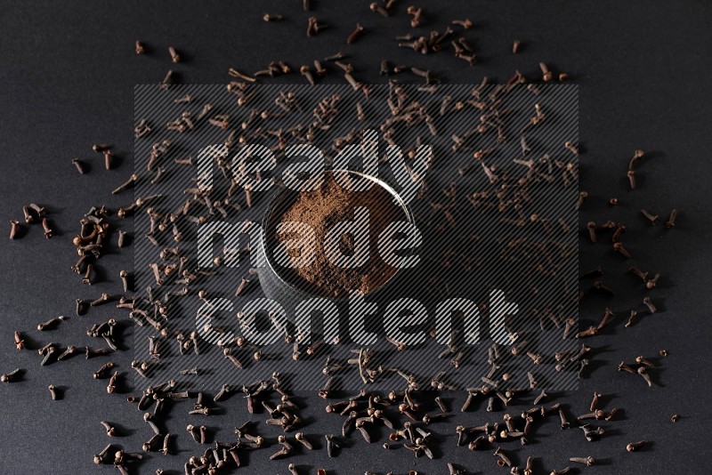 A black pottery bowl full of cloves powder and cloves whole grains on a black flooring