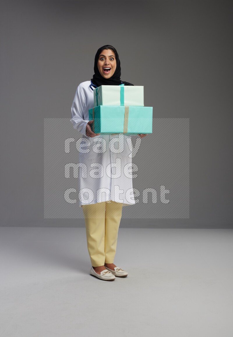 Saudi woman wearing lab coat with stethoscope standing holding gift box on Gray background
