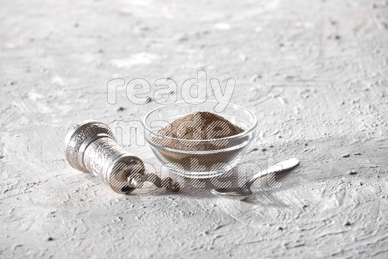A glass bowl full of black pepper powder and a metal spoon and a turkish metal grinder on textured white flooring