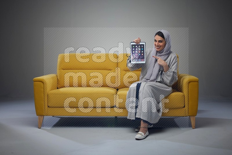 A Saudi female wearing a light gray Abaya and head scarf sitting on a yellow sofa and showing her tablet's screen while wearing headphones eye level on a grey background