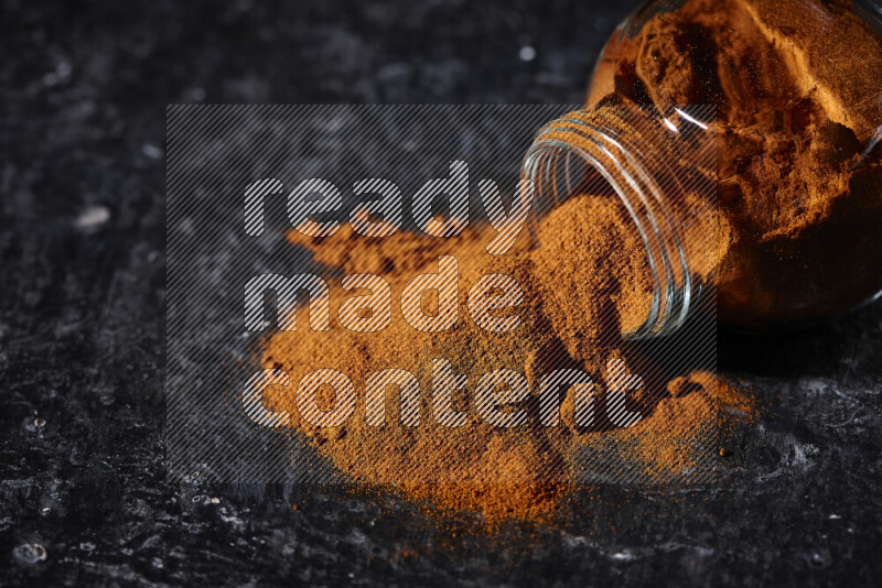 A glass jar full of ground paprika powder flipped with some spilling powder on black background