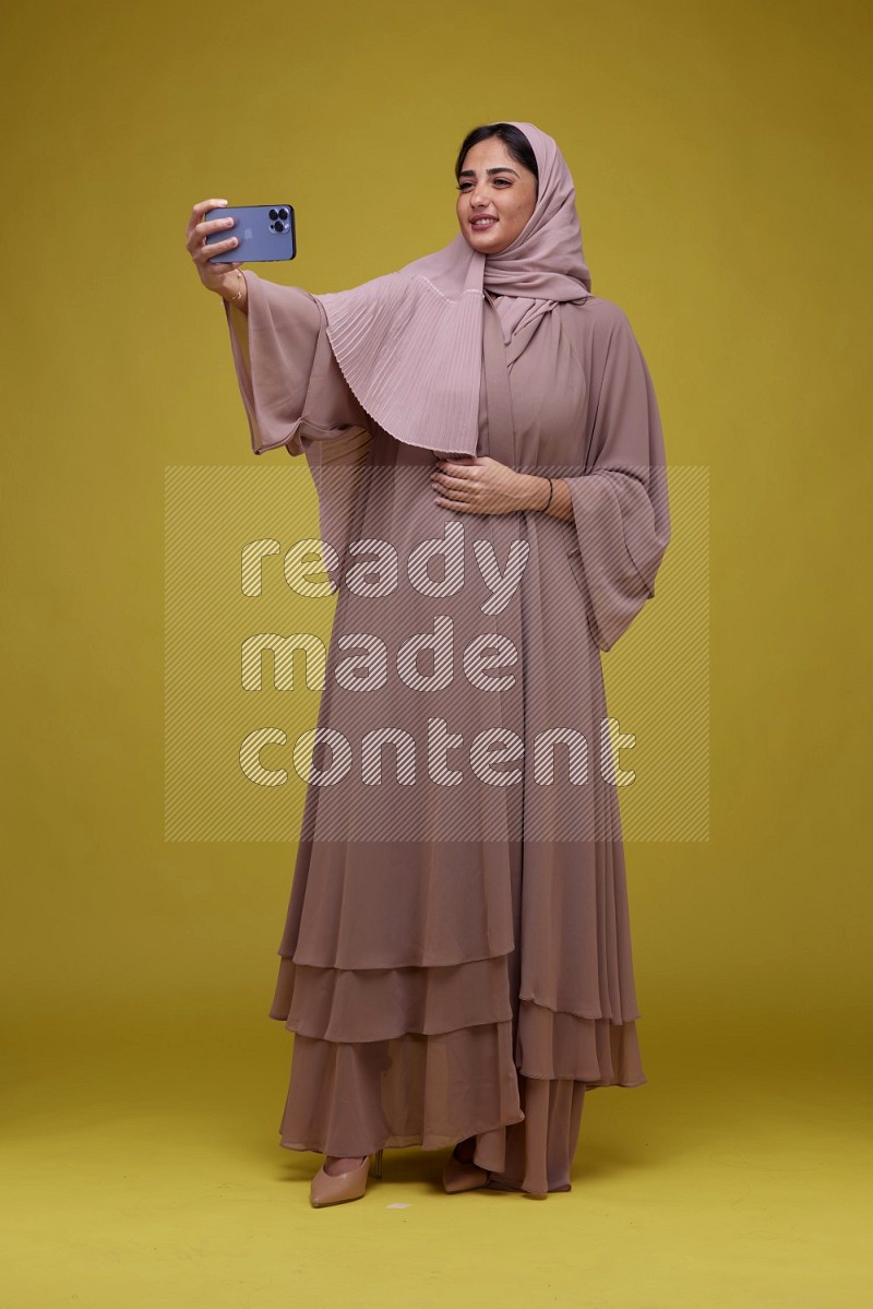 A woman taking a Selfie on a Yellow Background wearing Brown Abaya with Hijab
