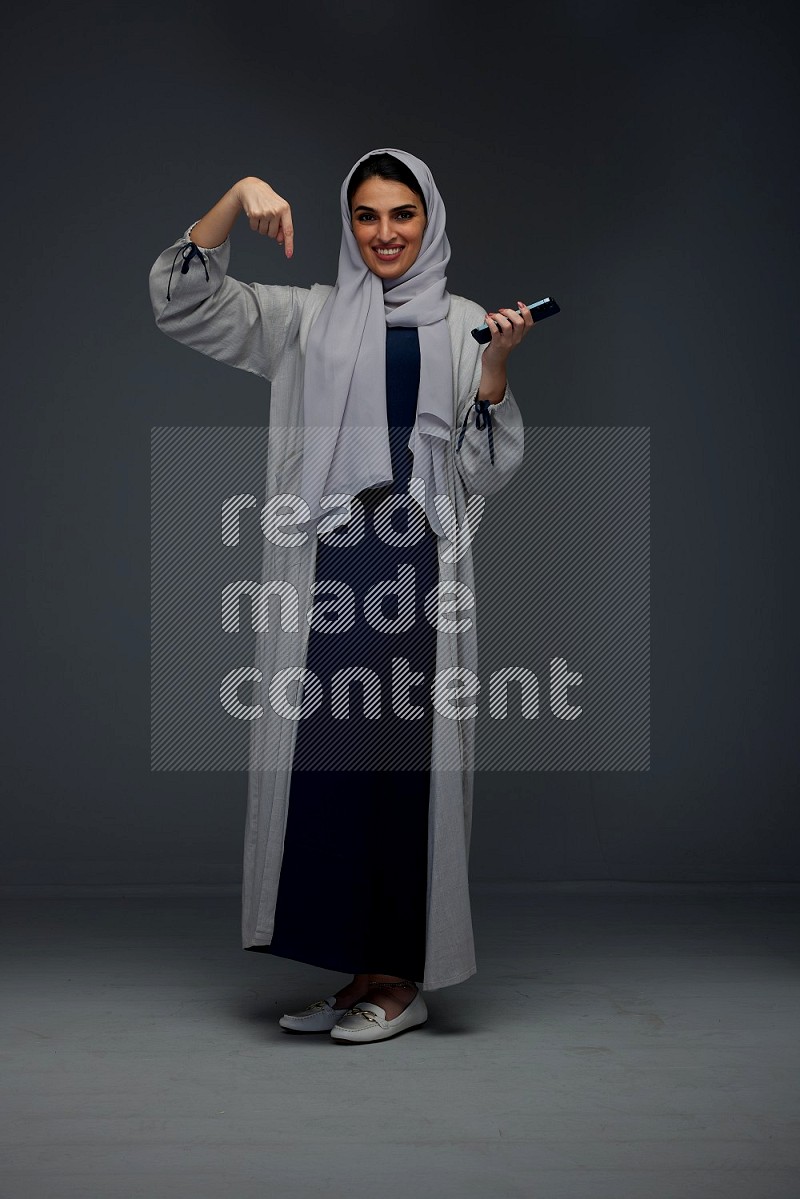 A Saudi woman wearing a light gray Abaya and head scarf standing and holding a phone on a grey background