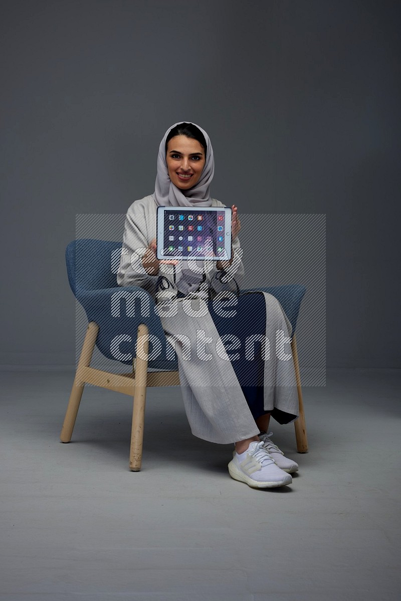 A Saudi woman wearing a light gray Abaya and head scarf sitting on a dark grey chair and showing the tablet's screen eye level on a grey background