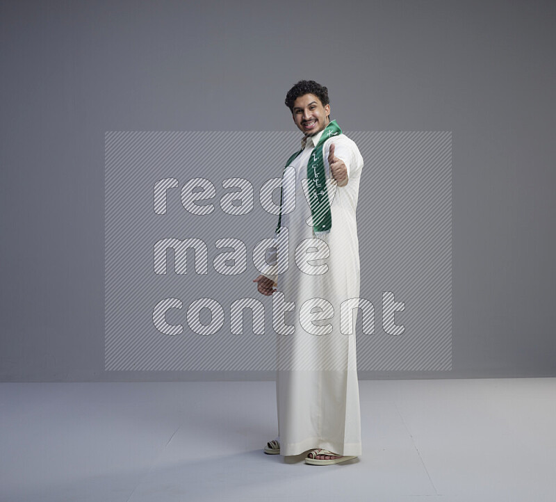 A Saudi man standing wearing thob and Saudi flag scarf on gray background